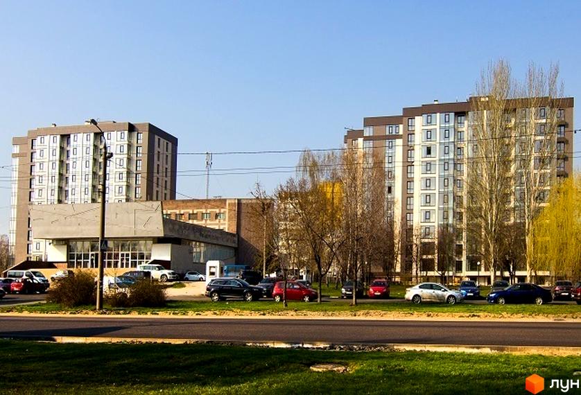 Житловий комплекс Салют - сучасні багатоповерхові будинки з великою кількістю вікон. На прибудинковій території розташовані дерева та парковка з автомобілями.