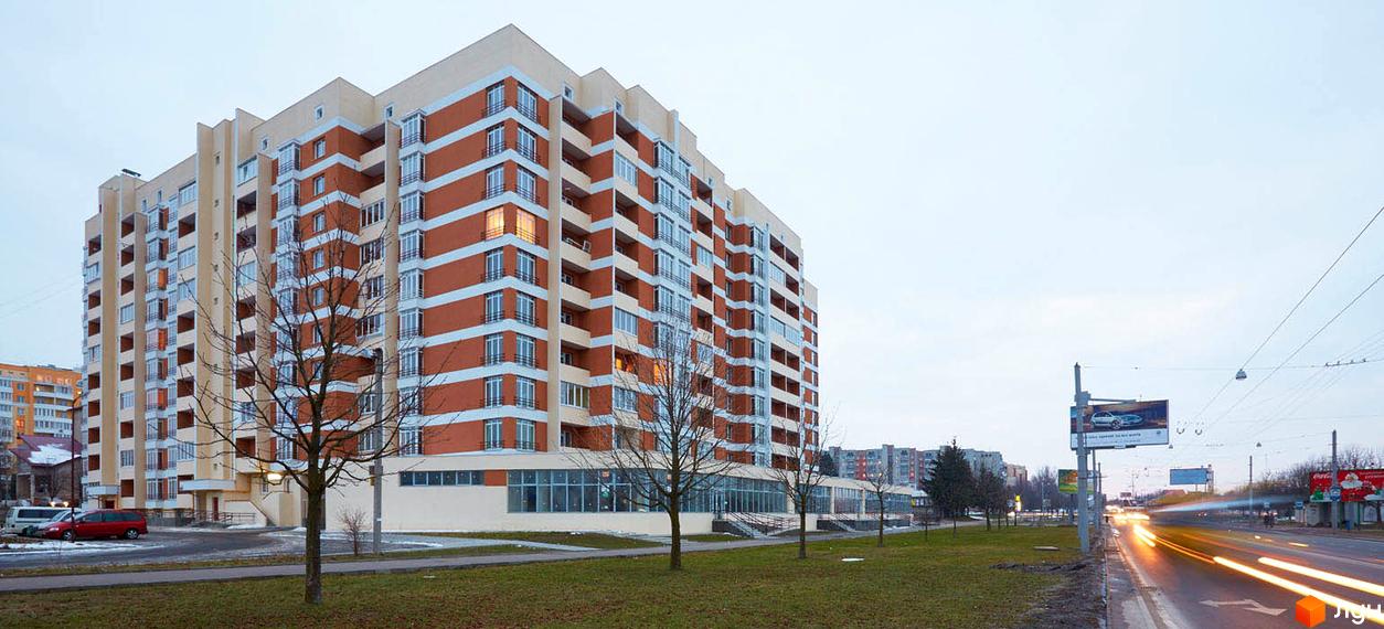 Зовнішній вигляд новобудови на вул. Перфецького, 2а, з цегляним фасадом та балконами. Навколо будинку зелена зона та дорога з автомобілями.