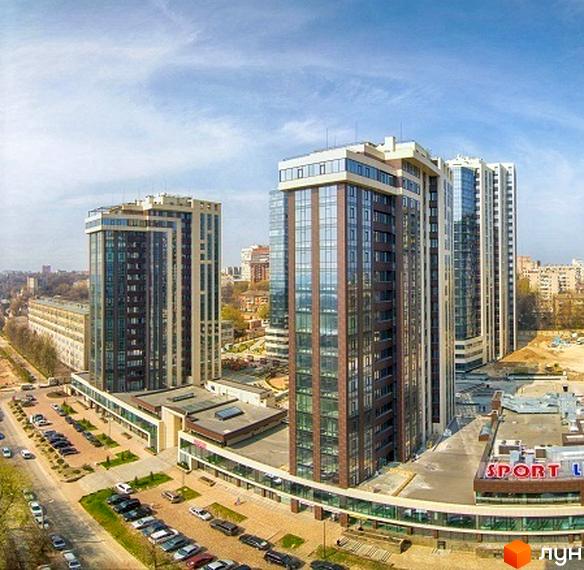 Високі будинки житлового комплексу Панорама з великими скляними вікнами. На прибудинковій території розташована парковка для автомобілів.