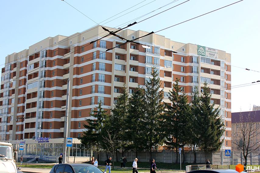 Багатоповерховий житловий комплекс на вул. Перфецького, 2а, з цегляним фасадом та великими вікнами. Навколо будинку ростуть зелені ялини, а перед ним розташована прибудинкова територія з деревами та огорожею.