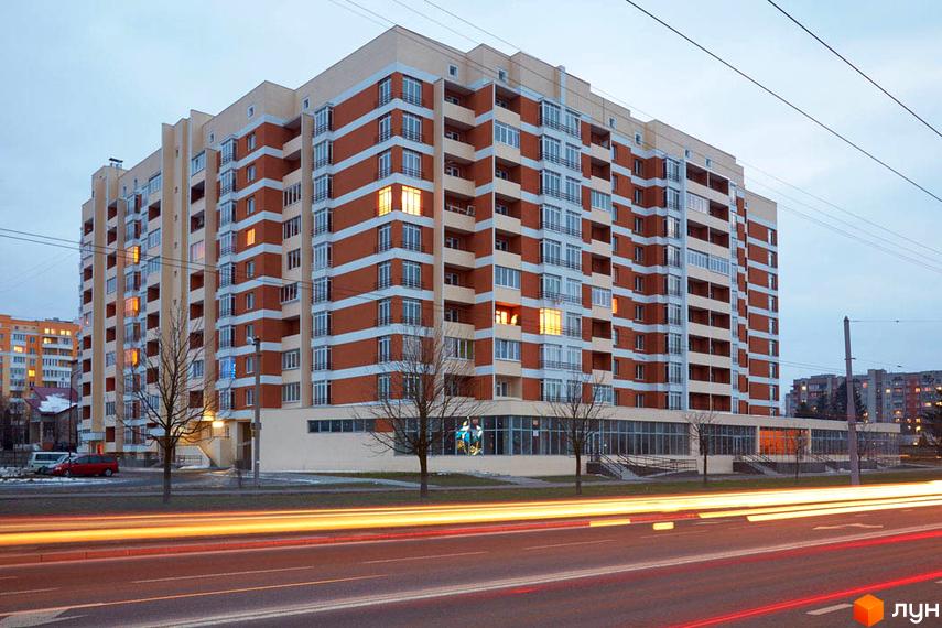 Фасад житлового комплексу на вул. Перфецького, 2а, з коричневими та білими смугами, балконами та великими вікнами. Поруч дорога з автомобільними світловими слідами.