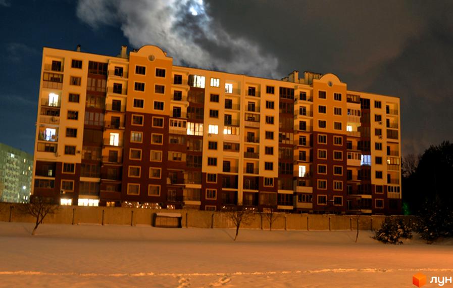 Житловий комплекс Паркове містечко вночі. Багатоповерховий будинок з жовтими та бордовими фасадами, освітленими вікнами. Навколо засніжена територія з деревами та парканом.