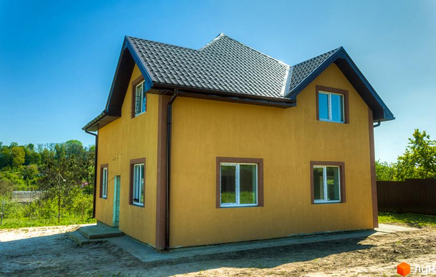 Двоповерховий будинок з жовтою штукатуркою та темним дахом у містечку котеджів Sunhills. Навколо будинку видно зелену територію та паркан.