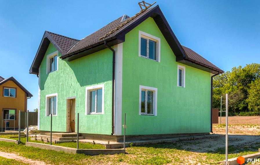 Зовнішній вигляд двоповерхового будинку з зеленими стінами та коричневим дахом у котеджному містечку КМ Sunhills. Навколо будинку облаштована прибудинкова територія з травою та бетонними елементами.