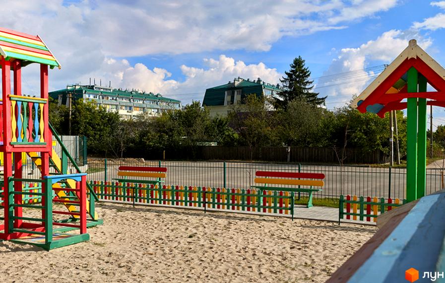 Дитячий майданчик з яскравим обладнанням та лавками на піщаному покритті. На задньому плані видно багатоповерхові будинки з зеленими дахами та дерева.