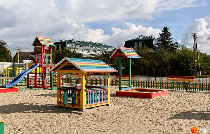 Дитячий майданчик з різнокольоровими гойдалками, гіркою та пісочницею. На задньому плані видно багатоповерхові будинки.