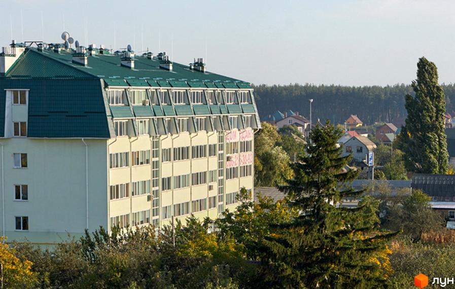 Зовнішній вигляд багатоквартирного будинку з зеленим дахом та безліччю вікон. Навколо будинку видно дерева та інші житлові будинки.