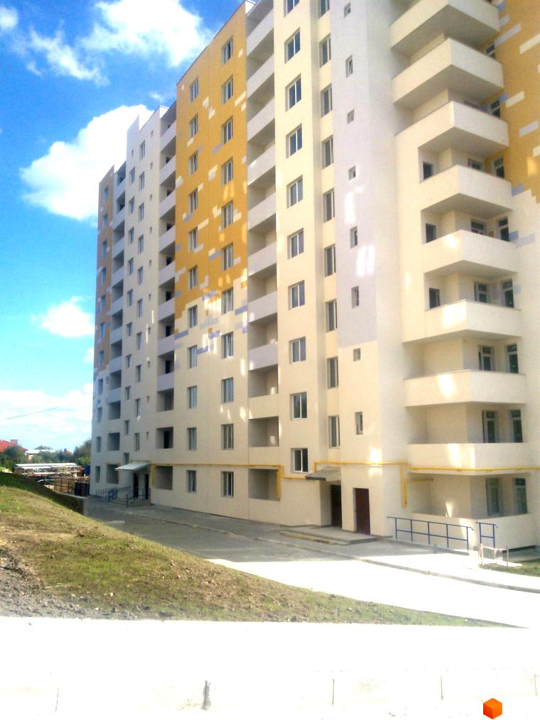 Зовнішній вигляд новобудови на вул. Під Голоском, 8. Багатоповерховий будинок з балконами та вікнами на тлі блакитного неба з хмарами. Прибудинкова територія з зеленим схилом та асфальтованою доріжкою.