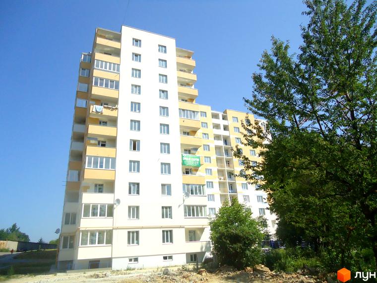 Зовнішній вигляд новобудови на вул. Під Голоском, 8. Будинок має світлий фасад з жовтими елементами та балконами. Навколо зелені дерева та елементи прибудинкової території.