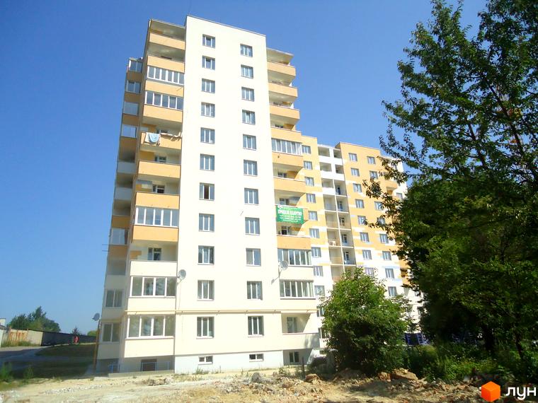 Новобудова на вул. Під Голоском, 8: високий будинок з жовтими та білими фасадами, балконами та великими вікнами. Навколо зелені дерева та ділянка з будівельним сміттям.