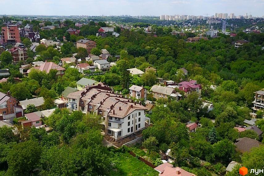 Зображення житлового комплексу з будинками з коричневими дахами та білими фасадами. Навколо будівель багато зелені та дерев.