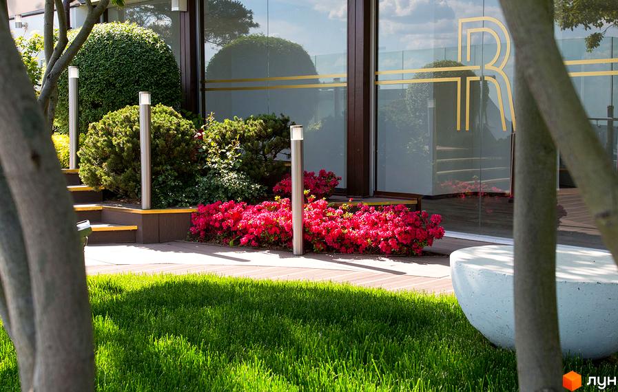 Житловий комплекс Royal Tower з великими скляними фасадами та логотипом R. Прибудинкова територія прикрашена яскравими квітами та зеленню.