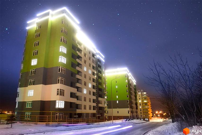 Житловий комплекс Пасічний у вечірній час з підсвічуванням фасаду. Новобудова з зеленими та сірими елементами на тлі зоряного неба та зимової дороги.