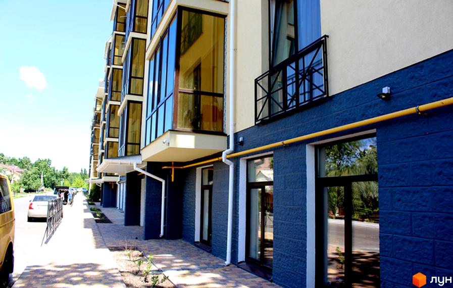 Зовнішній вигляд житлового комплексу Almond apartments з великими вікнами та темною цегляною обробкою. Поруч розташована доглянута прибудинкова територія з тротуаром та зеленими насадженнями.