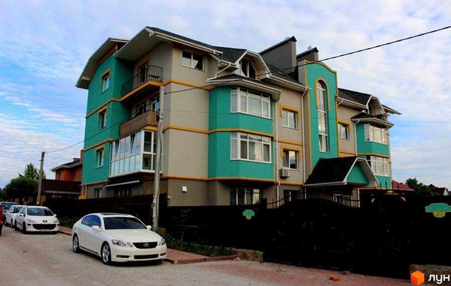 Багатоповерховий будинок з білими вікнами та балконами, пофарбований у сірий та бірюзовий кольори з жовтими смугами. Перед будинком припарковані білі автомобілі, а територія огороджена чорним кованим парканом.