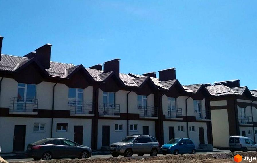 Таунхауси СанЛенд – це сучасні двоповерхові будинки з коричневими дахами та світлими фасадами. На території комплексу розташовані автомобілі.