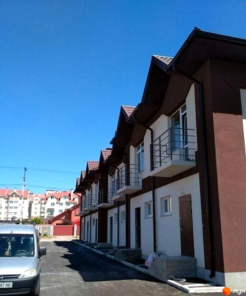 Зовнішній вигляд таунхаусів СанЛенд з коричневими фасадами та білими стінами, з балконами та вікнами. Поряд розташована прибудинкова територія з паркуванням та дорогою.