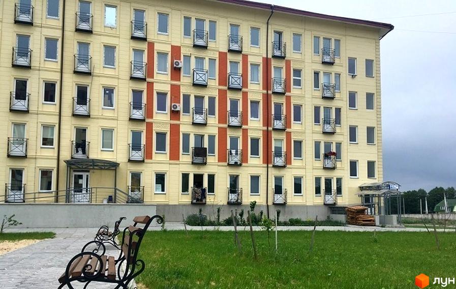 Фасад жовтого житлового комплексу Кошовий з червоними вставками та балконами. На прибудинковій території зелена галявина та лавка.