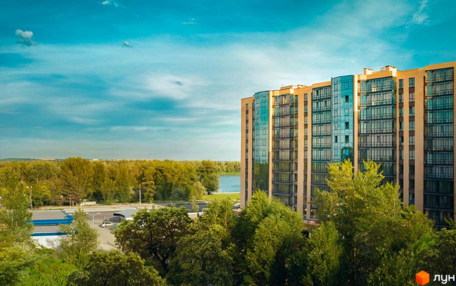 Багатоповерховий будинок Житлового комплексу River Park з бежевим фасадом та блакитним склінням. Навколо зелені дерева та видно річку.