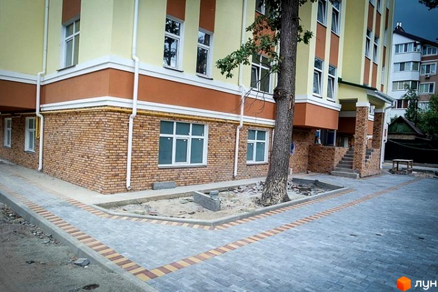 Зовнішній вигляд новобудови на вул. Шевченка, 22б, з цегляним цоколем та жовтими стінами. Прибудинкова територія з бруківкою та деревом біля входу.