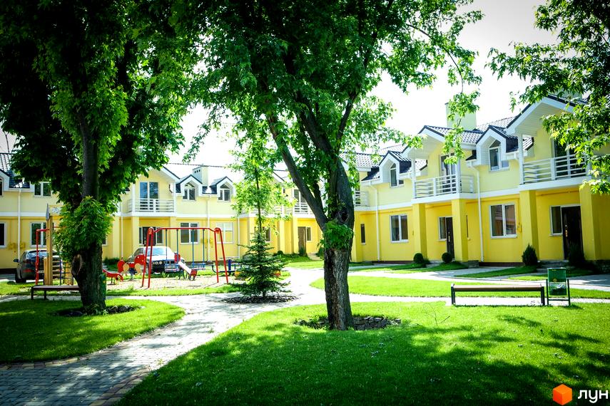 Котеджне містечко Sun City з жовтими будинками, білими балконами та чорними дахами. На території є зелений газон, дерева, лавки та дитячий майданчик.