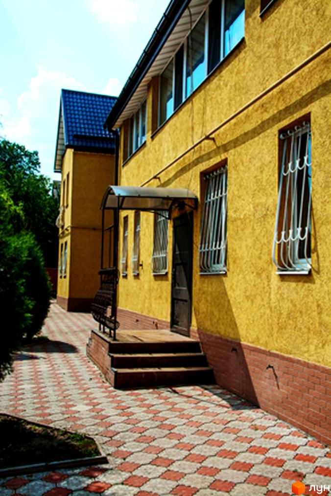 Жовтий двоповерховий будинок з вікнами під решітками та навісом над входом. Поруч доріжка з червоно-білої плитки та зелені.
