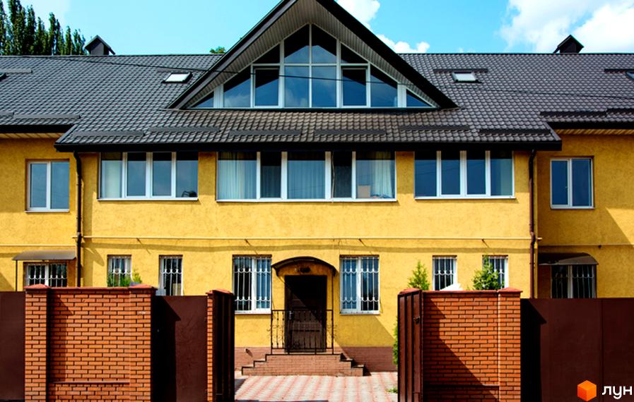 Таунхаус Санхауз з жовтим фасадом та темним дахом. Навколо будинку цегляний паркан та зелені насадження.