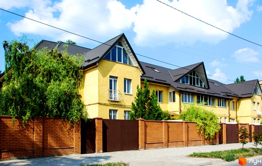 Жовтий двоповерховий будинок з темним дахом та великими вікнами. Огороджений цегляним парканом з коричневими воротами. Навколо багато зелені.