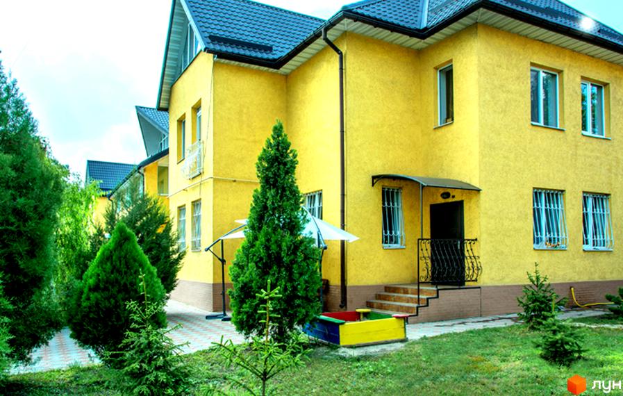 Житловий комплекс Санхауз: двоповерхова жовта будівля з темним дахом та зеленими деревами навколо. На прибудинковій території розташовані дитячий майданчик та доріжки.