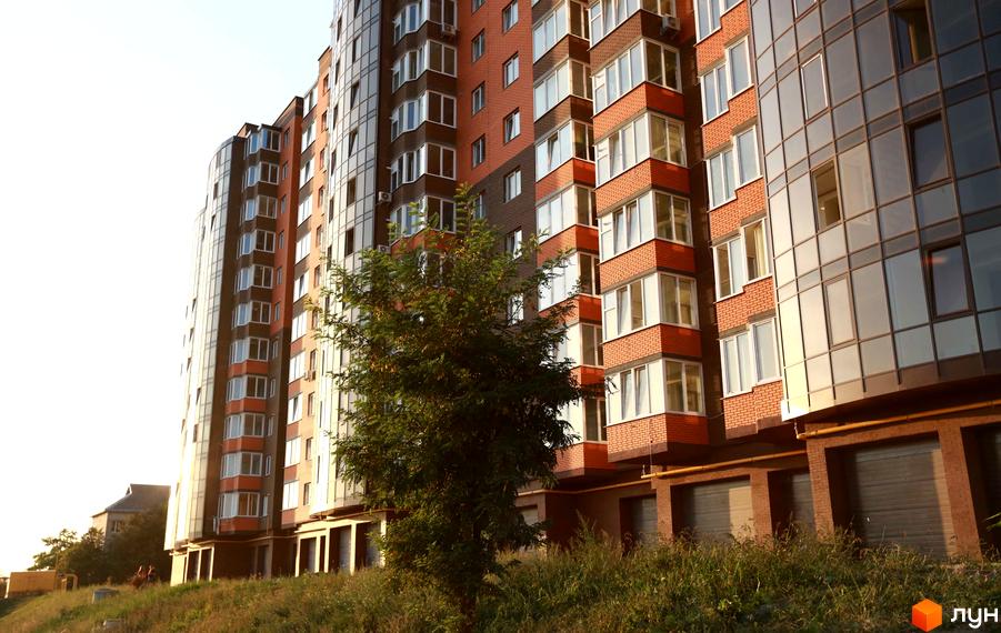 Багатоповерховий будинок Житлового комплексу Галицький Лев з цегляними та скляними фасадами, вікнами та балконами. Перед будинком зелений схил.