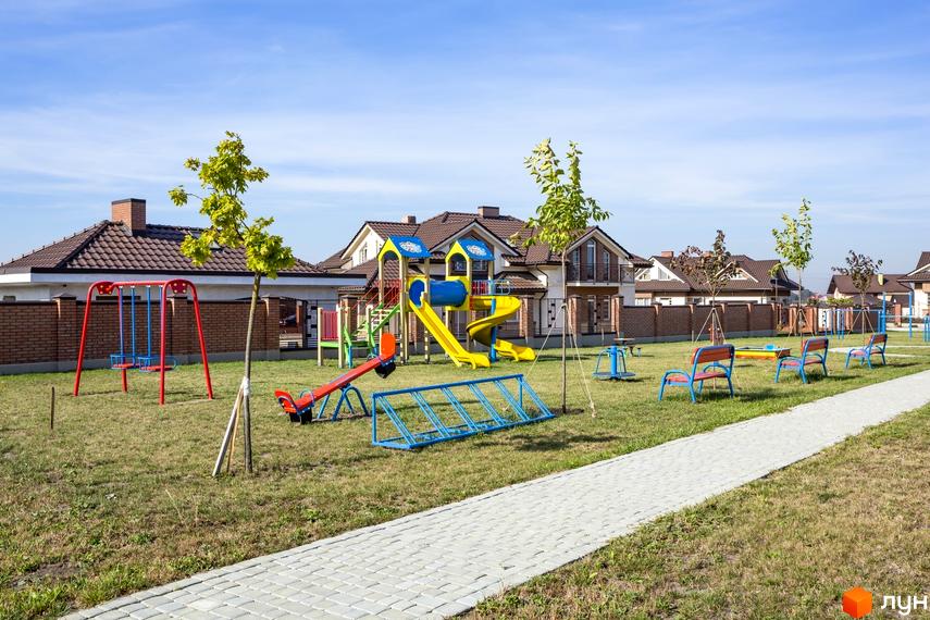 Котеджне містечко Панська Гора: дитячий майданчик з гірками, гойдалками, каруселлю та лавками на тлі двоповерхових будинків з коричневими дахами.