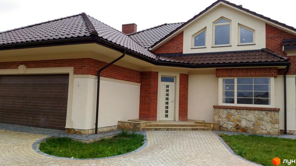 Котеджне містечко КМ Панська Гора: фасад будинку з цегляним оздобленням, великими вікнами та черепичним дахом. Прибудинкова територія з бруківкою та зеленню.