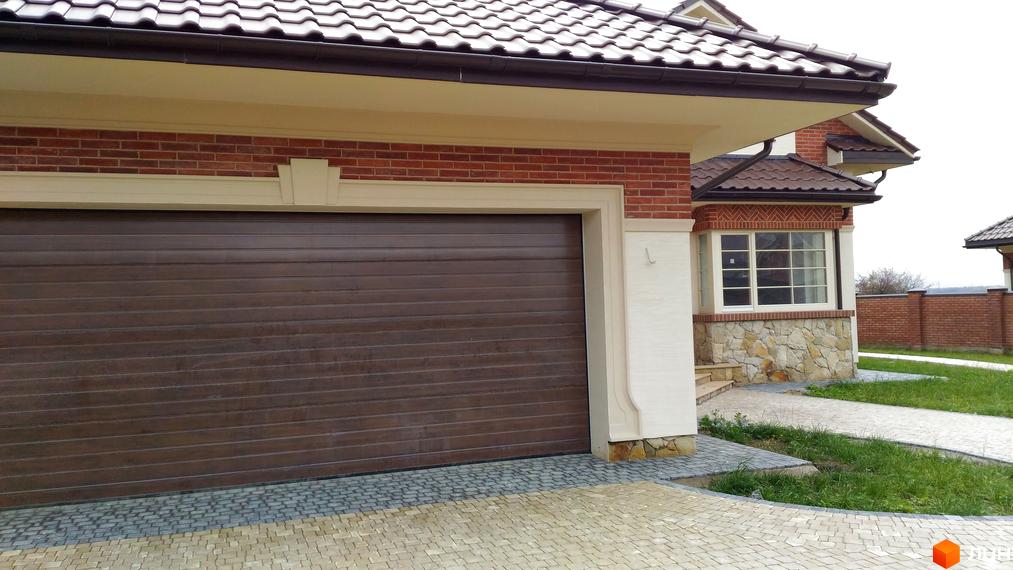 Зовнішній вигляд будинку з цегляним фасадом та гаражними воротами. Прибудинкова територія з бруківкою та зеленою травою.