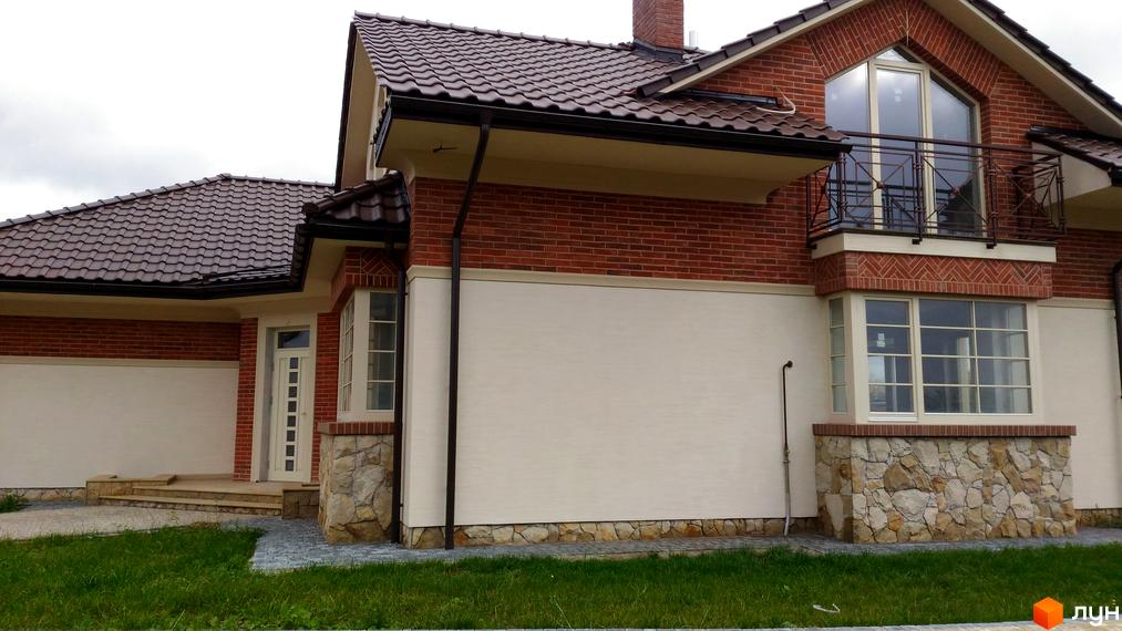 Зовнішній вигляд котеджного містечка КМ Панська Гора: цегляний будинок з коричневим черепичним дахом, світлими стінами та кам'яним цоколем. Навколо будинку зелена галявина та бруківка.