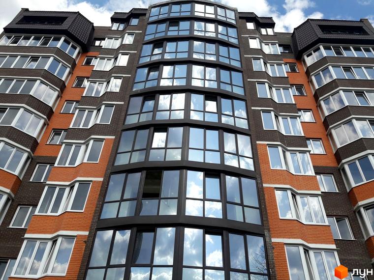 Фасад житлового комплексу Затишок з цегляними та скляними елементами. Багатоповерхова новобудова з великими вікнами, що відбивають хмари.