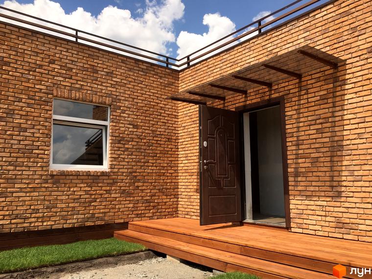 Зовнішній вигляд будинку з коричневої цегли з відкритими дверима. Перед будинком розташовані дерев'яні сходи та газон.