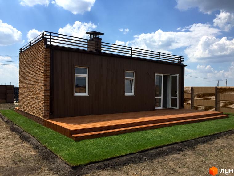 Одноповерховий будинок з коричневим фасадом та терасою з дерева. Поряд газон та доглянута прибудинкова територія. Котеджний комплекс „Фортеця Колонщини“.