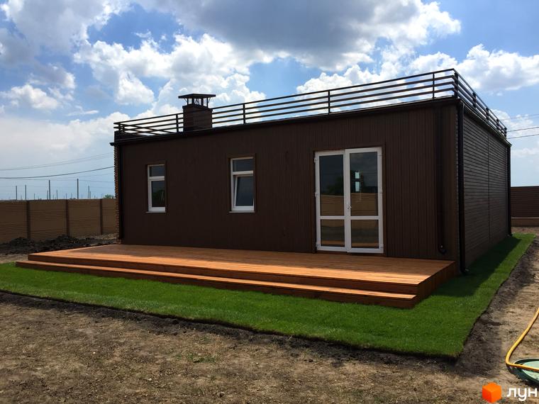 Одноповерховий будинок з дерев'яною терасою та газоном. Будівля має коричневі стіни, вікна та двері з білими рамами. На даху встановлено металеву огорожу.