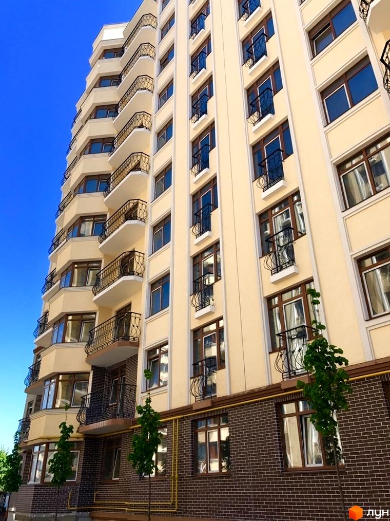 Новобудова Rich Town: висотна будівля з кремовим фасадом, балконами та цегляною основою. Зелені дерева біля входу.