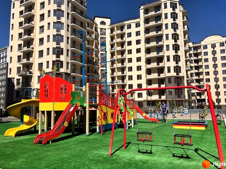Житловий комплекс Rich Town: фасади світло-бежевих багатоповерхових будинків та дитячий майданчик з гірками та гойдалками.