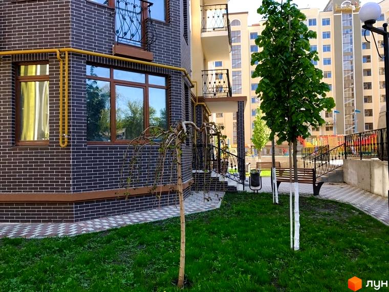 Зовнішній вигляд новобудови ЖК Rich Town з темної цегли та вікнами з коричневими рамами. На прибудинковій території зелений газон, дерева та лавки.