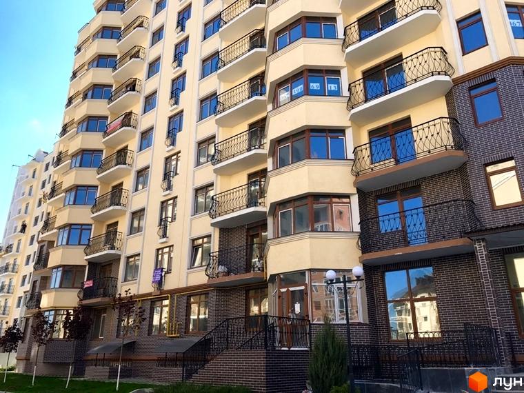 Зовнішній вигляд багатоповерхового житлового комплексу Rich Town з балконами та цегляним оздобленням. Навколо будинку зелені насадження та доглянута територія.