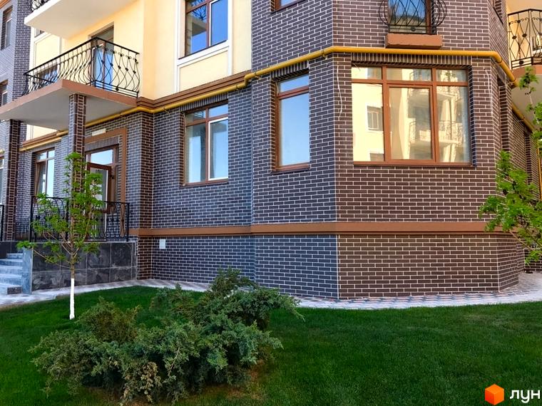 Фасад новобудови ЖК Rich Town з коричневої цегли та великими вікнами. Поруч доглянутий газон та зелені кущі.