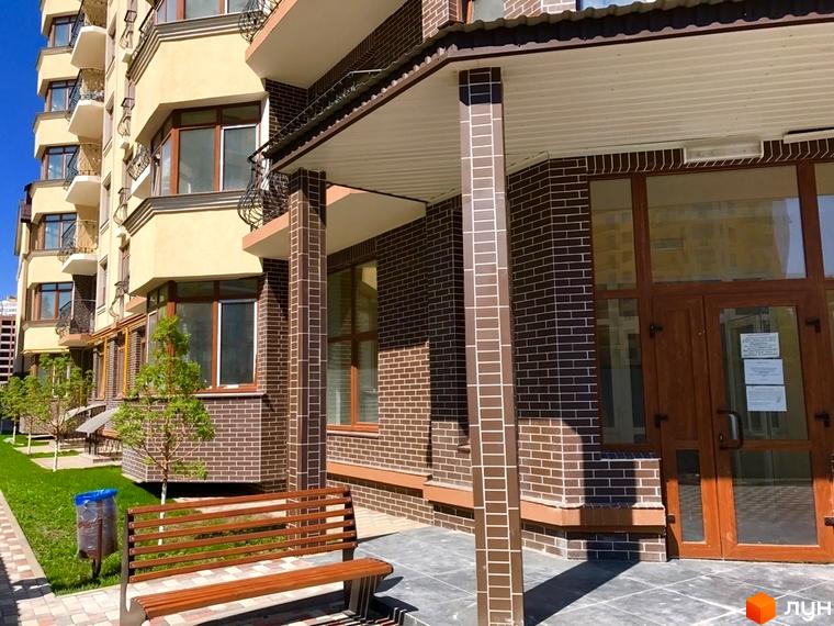 Зовнішній вигляд новобудови ЖК Rich Town з цегляним фасадом та дерев'яними дверима. Прибудинкова територія з лавкою та зеленим газоном.