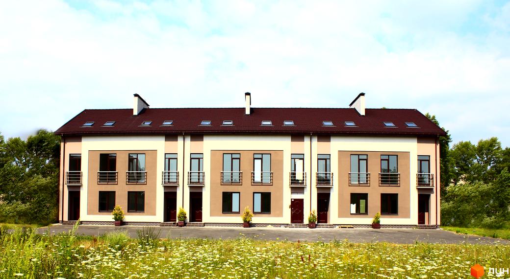 Ряд таунхаусів Деснянська садиба з бежево-коричневим фасадом, темним дахом та мансардними вікнами. Перед будинками поле з польовими квітами та зелені дерева на фоні.