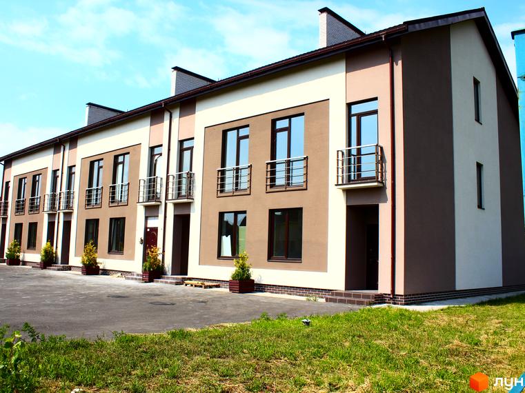 Таунхаус „Деснянська садиба“: ряд двоповерхових таунхаусів коричневого та бежевого кольорів з балконами. Перед будинками асфальтований майданчик та зелена трава.