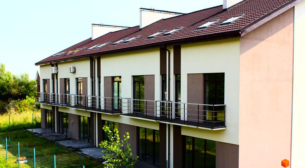 Таунхаус „Деснянська садиба“: світлий фасад з коричневими вставками, балкони з поручнями та дах з черепиці з мансардними вікнами. Прибудинкова територія облаштована газоном.