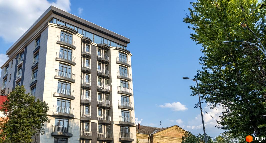 Клубний будинок Tarasovskiy Apartments має світлий фасад з темними акцентами та численними балконами. Навколо будинку видно зелені дерева та елементи міського освітлення.