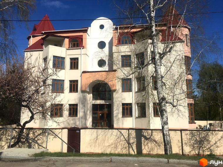 Фасад Клубного будинку „Статус“ з світлими стінами, червоними дахами, круглими вікнами та арковим входом. Територія має дерева та огорожу.