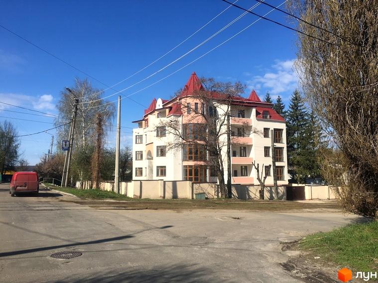 Клубний будинок „Статус“: багатоповерхова будівля зі світлим фасадом, червоним дахом та балконами. На передньому плані дорога, дерева та паркан.